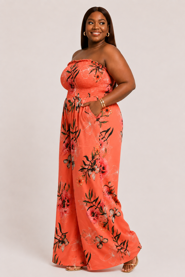 A woman modeling a strapless coral-orange maxi dress with a black and gray floral print, standing against a light gray studio background.