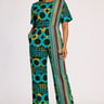 Woman wearing a teal and black Ankara circle print wide leg jumpsuit with a diagonal woven Aso Oke stripe panel in teal gold and grey running from shoulder to hem styled with gold strappy heeled sandals against a white background