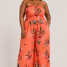A woman modeling a strapless wide-leg jumpsuit in a bright coral-orange color with a black and red floral print, set against a plain light gray studio background.