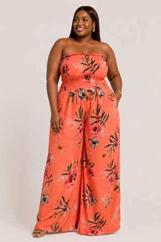 A woman modeling a strapless wide-leg jumpsuit in a bright coral-orange color with a black and red floral print, set against a plain light gray studio background.