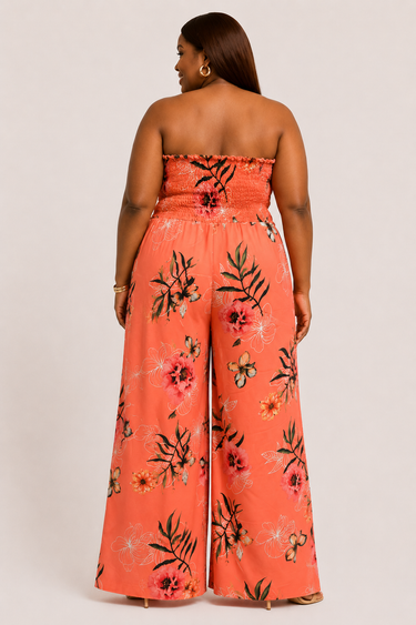 A woman shown from the back wearing a strapless wide-leg jumpsuit in a bright coral-orange color with a black and pink floral print, set against a plain light gray studio background.