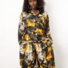 A woman with curly black hair stands against a plain white studio background wearing a matching long-sleeve button-front top and wide-leg pants set in an abstract print of black, yellow, orange, and off-white.
