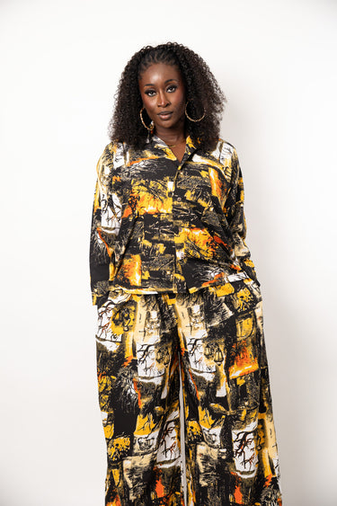 A woman with curly black hair stands against a plain white studio background wearing a matching long-sleeve button-front top and wide-leg pants set in an abstract print of black, yellow, orange, and off-white.