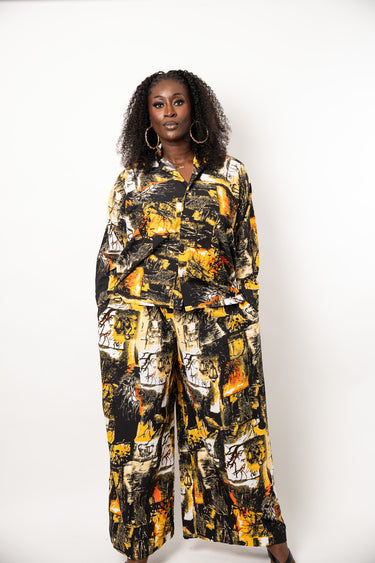 A woman modeling a matching two-piece jumpsuit set with a bold abstract print in black, yellow, orange, and white, standing against a plain white studio background.