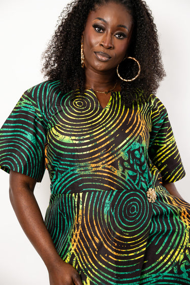 A woman wearing a short-sleeve V-neck dress with a vibrant green, yellow, and black spiral pattern, styled with gold hoop earrings against a plain white background.
