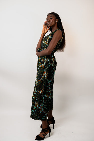 A woman modeling a sleeveless black maxi dress with a green and teal abstract botanical pattern, paired with black strappy high heels, against a plain white studio background.