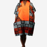 A woman modeling a short, loose V-neck dress with a bright orange center panel and a geometric patchwork pattern in gray, rust red, black, and white on the sleeves and lower skirt, photographed against a light gray studio background.