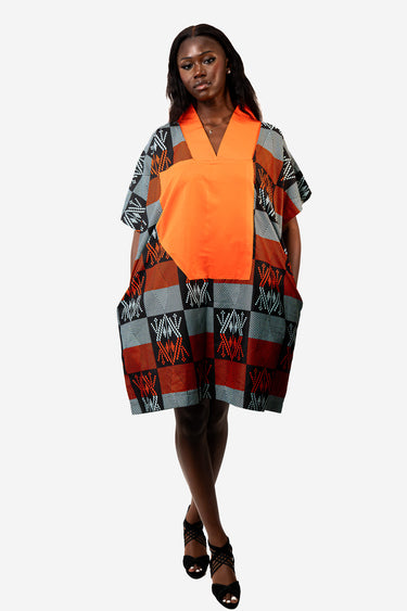 A woman modeling a short, loose V-neck dress with a bright orange center panel and a geometric patchwork pattern in gray, rust red, black, and white on the sleeves and lower skirt, photographed against a light gray studio background.