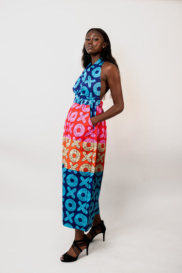 A woman modeling a sleeveless halter maxi dress with a vibrant multicolor patchwork print featuring blue, pink, orange, and yellow patterns, paired with black strappy high-heeled sandals, against a plain white studio background.