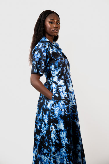 A woman modeling a short-sleeve collared dress with a blue and black tie-dye pattern, photographed against a plain light gray studio background.