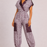 A woman modeling a short-sleeve jumpsuit with a purple and white patterned design, dark purple contrast trim, and cargo pockets, set against a solid light gray background.
