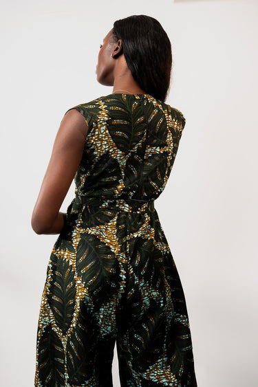 A woman shown from the back wearing a sleeveless jumpsuit with a dark green and black tropical leaf print accented with gold and teal details, set against a plain white studio background.