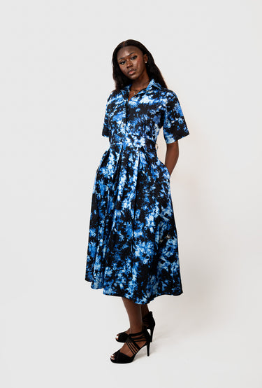 A woman modeling a short-sleeve midi shirt dress with a blue and black abstract tie-dye pattern, paired with black strappy heels, against a plain light gray studio background.