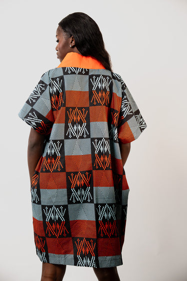 A woman shown from the back wearing a short-sleeve kaftan dress with a bold geometric pattern in orange, red, gray, black, and white, featuring a solid bright orange collar, set against a plain light gray background.