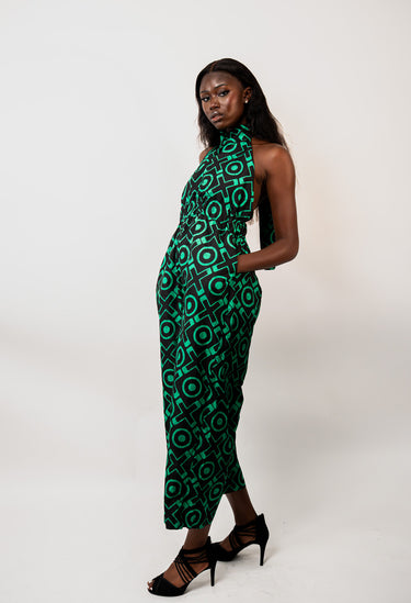 A woman modeling a sleeveless high-neck midi dress with a bold green and black geometric circular pattern, paired with black strappy high-heeled sandals, set against a plain light gray studio background.