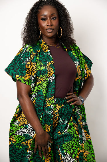 A woman modeling a matching two-piece outfit featuring a green, yellow, and white floral print wrap jacket and trousers, worn over a solid dark brown top, against a plain light background.