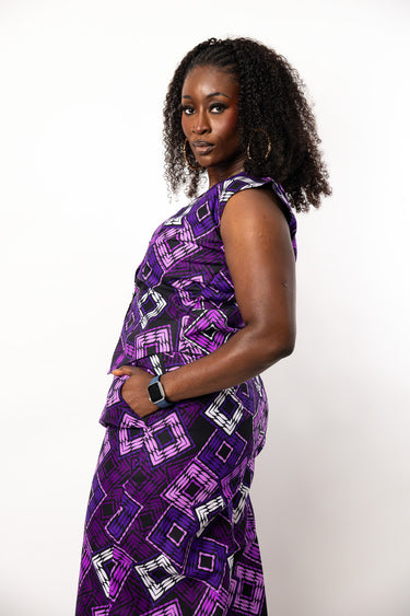A woman with curly black hair is shown from the waist up, wearing a sleeveless purple and black geometric print dress with white accents. She is standing against a plain white studio background, looking over her shoulder.