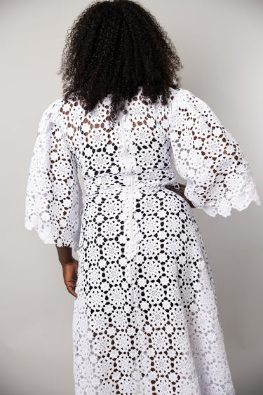 A woman viewed from behind wearing a white crochet lace dress with voluminous elbow-length sleeves and a matching waist tie, against a plain light gray studio background.