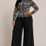 A woman modeling a long-sleeve mock-neck top with a black, white, and green geometric and floral pattern, paired with wide-leg black pants, against a solid light beige studio background.