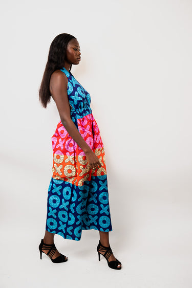 A woman modeling a sleeveless maxi dress with a vibrant, multi-colored African wax print pattern featuring blue, pink, orange, and yellow geometric and circular motifs, set against a plain white studio background.
