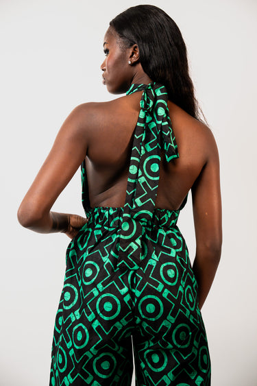 A woman shown from the back wearing a green and black geometric print halter jumpsuit against a plain light gray studio background.