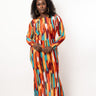 A woman modeling a long-sleeve maxi dress with a vibrant vertical ikat-style pattern in orange, red, yellow, blue, and white, standing against a plain white studio background.