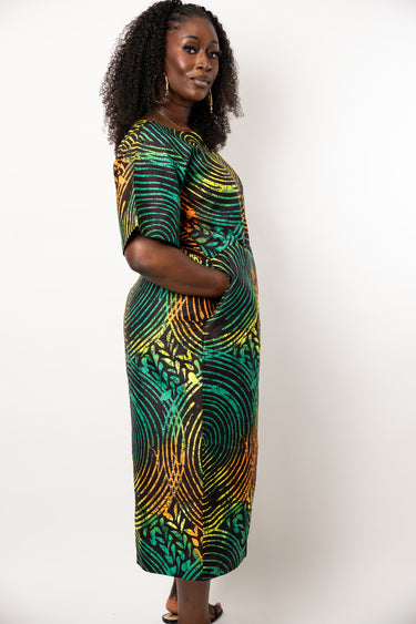 A woman modeling a short-sleeve midi dress with a vibrant green, yellow, and orange abstract pattern against a plain white studio background.
