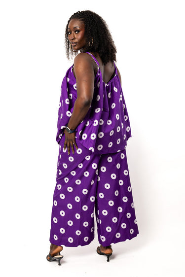 A woman modeling a purple sleeveless jumpsuit with a white circular pattern, photographed against a plain white studio background.