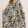 A woman modeling a loose, flowy kaftan dress with a bold abstract botanical pattern in black, white, and metallic gold, set against a plain light beige studio background.
