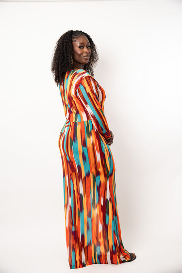 A woman with curly black hair stands in profile wearing a long-sleeved maxi dress with a vibrant vertical abstract pattern in orange, red, teal, and white, set against a plain white studio background.