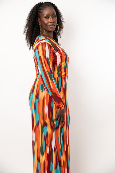 A woman with curly black hair stands in profile wearing a long-sleeve maxi dress with a vibrant vertical abstract pattern in orange, red, teal, and white, set against a plain off-white background.
