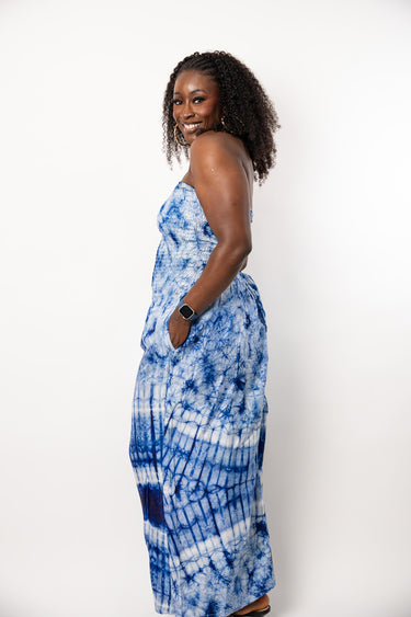 A woman with curly dark hair stands in profile wearing a strapless blue and white tie-dye maxi dress with pockets, set against a plain white studio background.