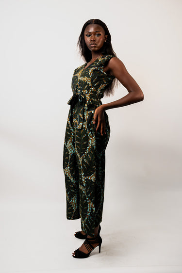 A woman modeling a dark green sleeveless jumpsuit with a yellow and white tropical floral pattern, paired with black strappy heels, against a plain white studio background.