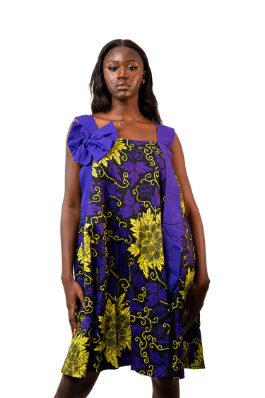 A woman modeling a sleeveless dress with a purple and yellow floral print, featuring a large purple bow detail on the left shoulder, set against a plain white background.