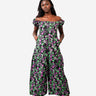 A model wearing a black off-the-shoulder jumpsuit with a vibrant pink and green floral print, styled with white sneakers against a solid light gray background.