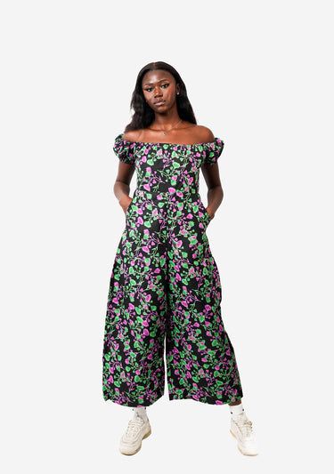A model wearing a black off-the-shoulder jumpsuit with a vibrant pink and green floral print, styled with white sneakers against a solid light gray background.