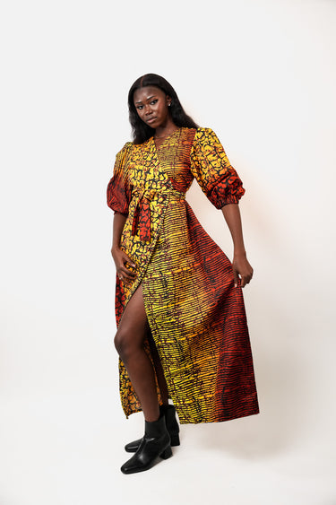 A woman modeling a long African wax print dress with puff sleeves and a high leg slit, featuring a vibrant yellow, red, and black pattern, set against a plain white studio background.