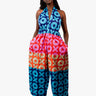 A woman modeling a sleeveless halter jumpsuit with a vibrant, multicolor African wax print featuring geometric patterns in blue, pink, orange, and red, paired with black strappy high-heeled sandals against a plain white studio background.