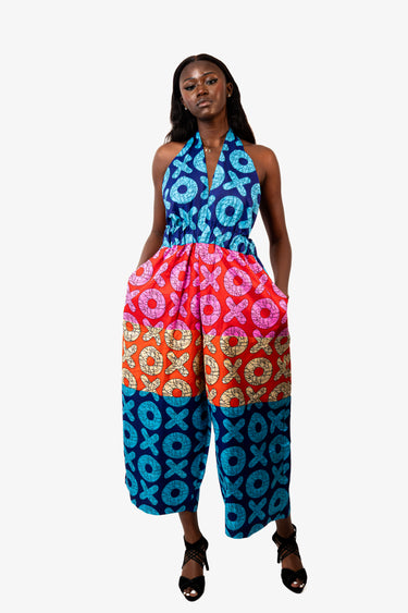 A woman modeling a sleeveless halter jumpsuit with a vibrant, multicolor African wax print featuring geometric patterns in blue, pink, orange, and red, paired with black strappy high-heeled sandals against a plain white studio background.