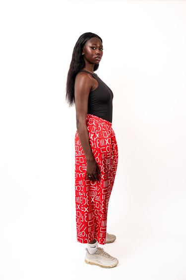 A woman stands in profile against a plain white studio background, wearing a black sleeveless top, red wide-leg pants with a white abstract print, white socks, and light beige sneakers.