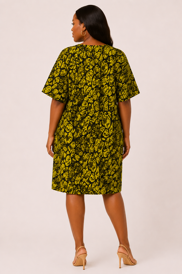 Plus size model wearing a bold olive green and black Ankara abstract print short shift dress with round scoop neckline wide kimono sleeves and relaxed fit styled with nude strappy heeled sandals against a light background