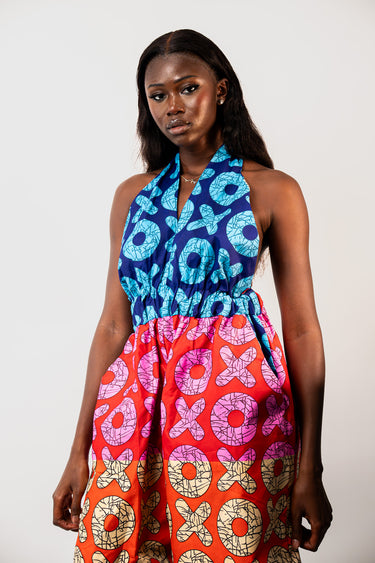 A model wearing a sleeveless halter dress with a vibrant, multi-colored African wax print. The dress features a blue and turquoise pattern on the upper bodice and a bright red and orange patterned skirt with pink, yellow, and beige motifs.